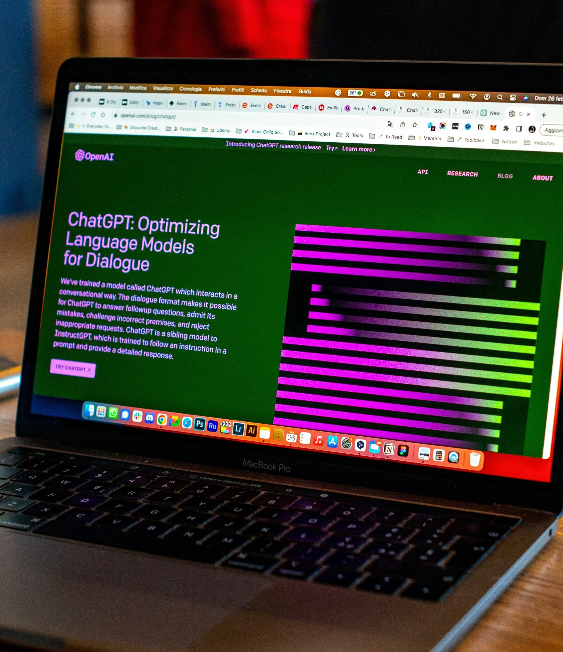 A laptop displaying the OpenAI website with a focus on ChatGPT. The screen shows a title "ChatGPT: Optimizing Language Models for Dialogue" alongside colorful graphics and text on a vibrant green background. The laptop is on a wooden table.