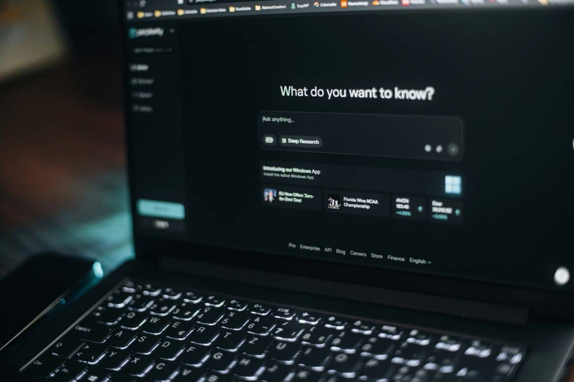 A laptop keyboard in focus with a blurred screen displaying a dark-themed search engine page asking, “What do you want to know?” and showing related research results below the search bar.