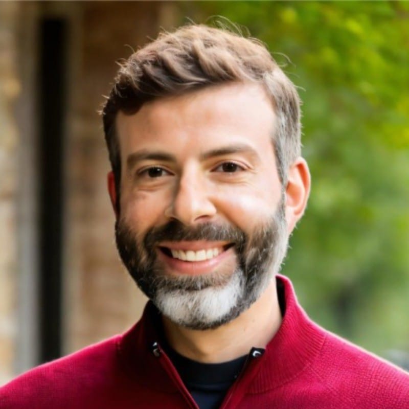 A man with short brown hair and a beard, wearing a maroon zip-up sweater, smiles at the camera outdoors with greenery and a brick wall in the blurred background.