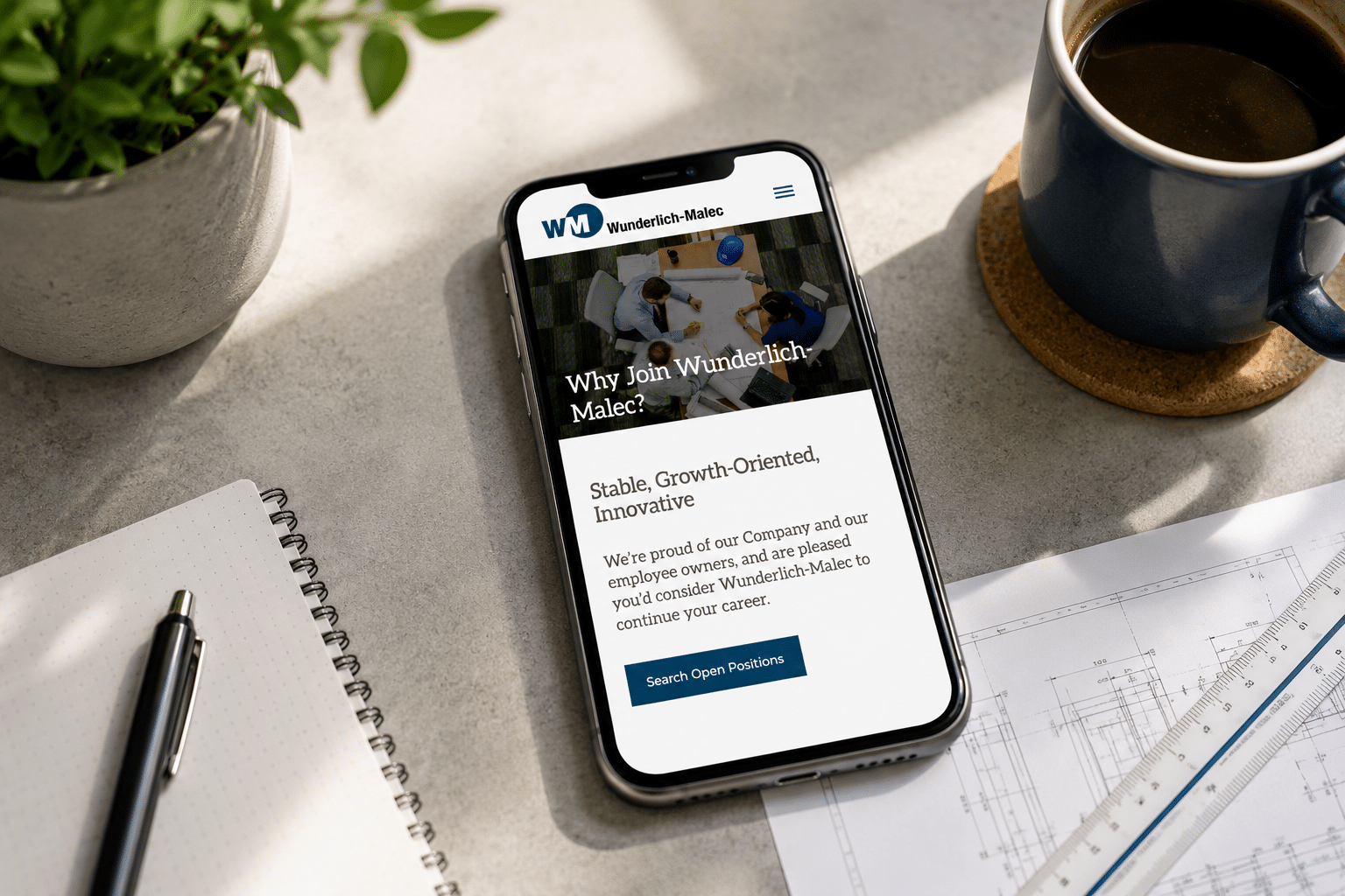 A smartphone displays a career page for Wunderlich-Malec on a desk with a notebook, pen, blue coffee mug, and architectural plans in sunlight.