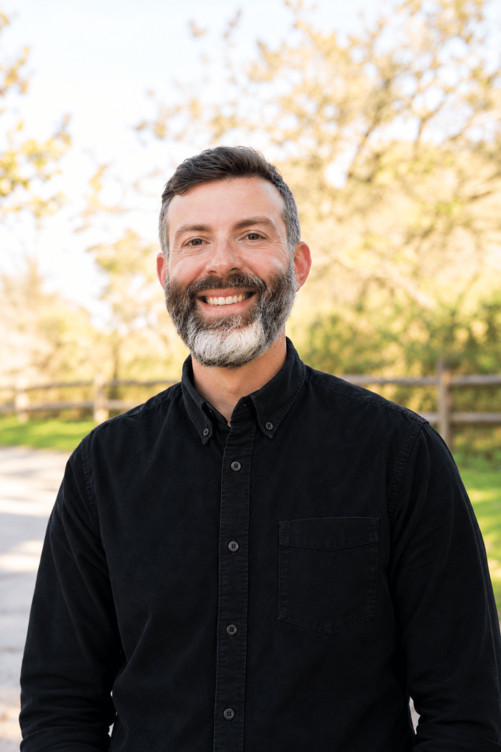 A smiling man with a beard wearing a black button-down shirt stands outdoors.